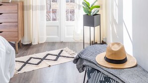 A room with a large window, a wooden dresser, a plant in a black pot, a hat on a bench, and a patterned rug on the floor. - MaisonMars La Maison de Famille (Marseille)