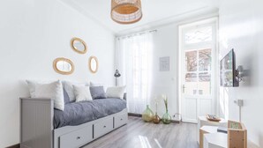 A modern bedroom with a grey bed, white walls, and a wooden floor. There are three decorative mirrors on the wall, a window with sheer curtains, and a television mounted on the wall. - MaisonMars La Maison de Famille (Marseille)