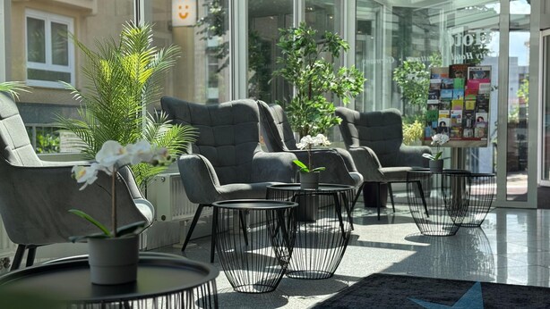 A modern indoor seating area with grey upholstered chairs, black wire tables, and potted plants. - Mirage City Hotel Stuttgart (Stuttgart)