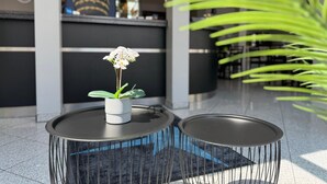 A reception area with a modern design, featuring a reception desk, a potted orchid, and two black tables with a unique wireframe design. - Mirage City Hotel Stuttgart (Stuttgart)