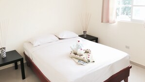 Una cama con sábanas blancas impecables, un control remoto y una toalla con motivos florales sobre un cabecero de madera. Hay dos mesitas de noche con jarrones decorativos y una ventana con cortina.