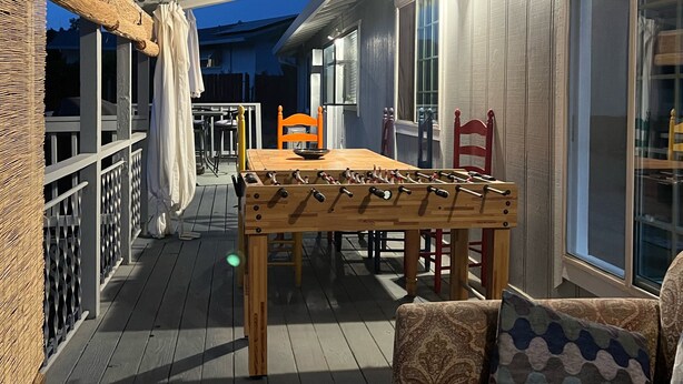 Um espaço ao ar livre coberto, com mesa e cadeiras de madeira, um sofá e uma janela com cortinas.