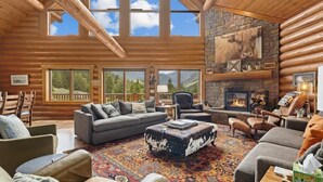 A spacious log cabin interior with a large fireplace, multiple seating areas, and large windows offering mountain views. - Last Resort (Red Lodge)