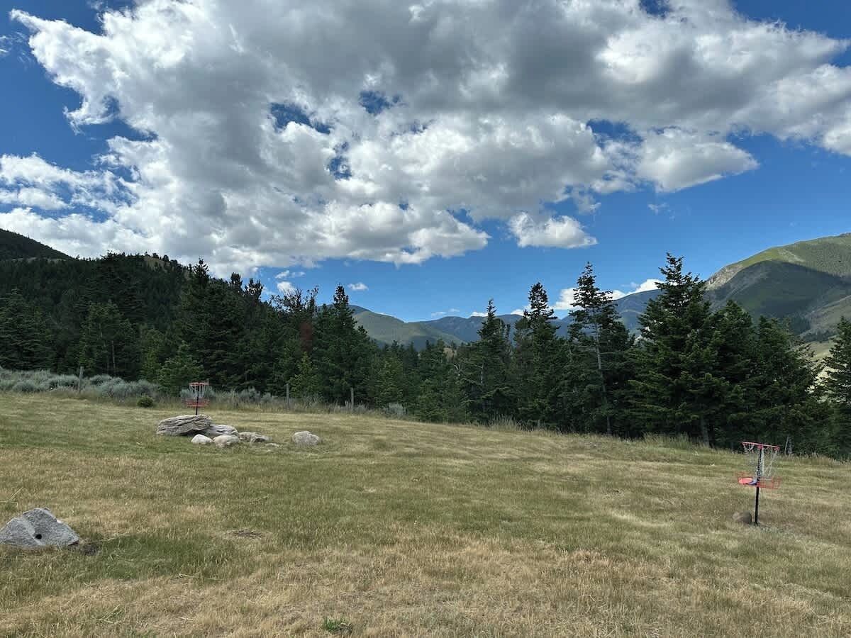 Un champ herbeux avec deux cibles pour lancer des frisbees, entouré d’arbres et de montagnes, sous un ciel partiellement nuageux.