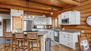 A modern kitchen with white cabinets, a stainless steel refrigerator, a microwave, and a large island with bar stools. - Last Resort (Red Lodge)