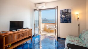 A room with a TV on a wooden cabinet, a balcony with a table and chairs, and a poster on the wall. - Holiday Home "Pergola 714" Steps from the Beach with Sea View and Balcony (STINTINO)