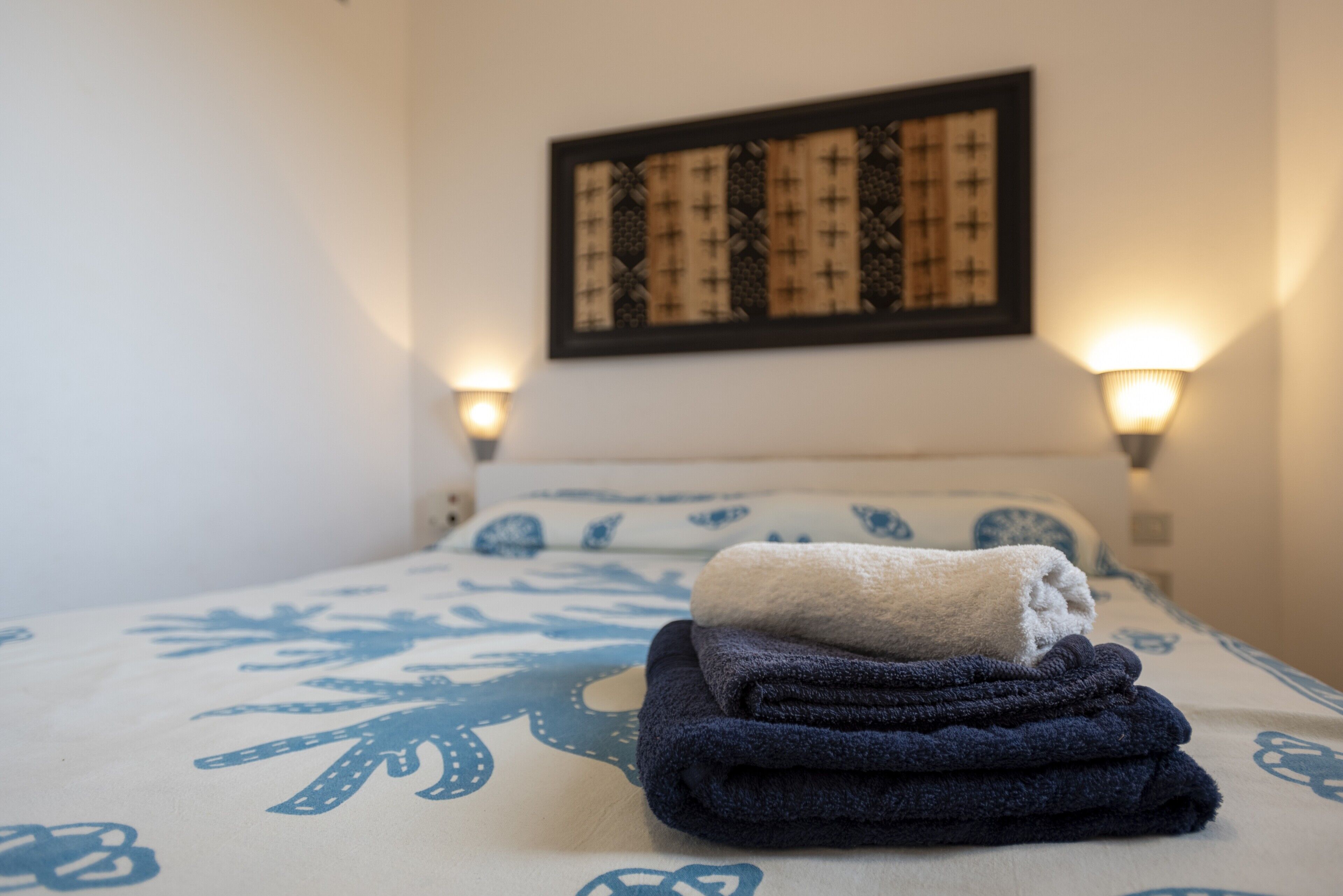 A neatly made bed with a patterned bedspread, two wall-mounted lamps, and a framed artwork above the bed.