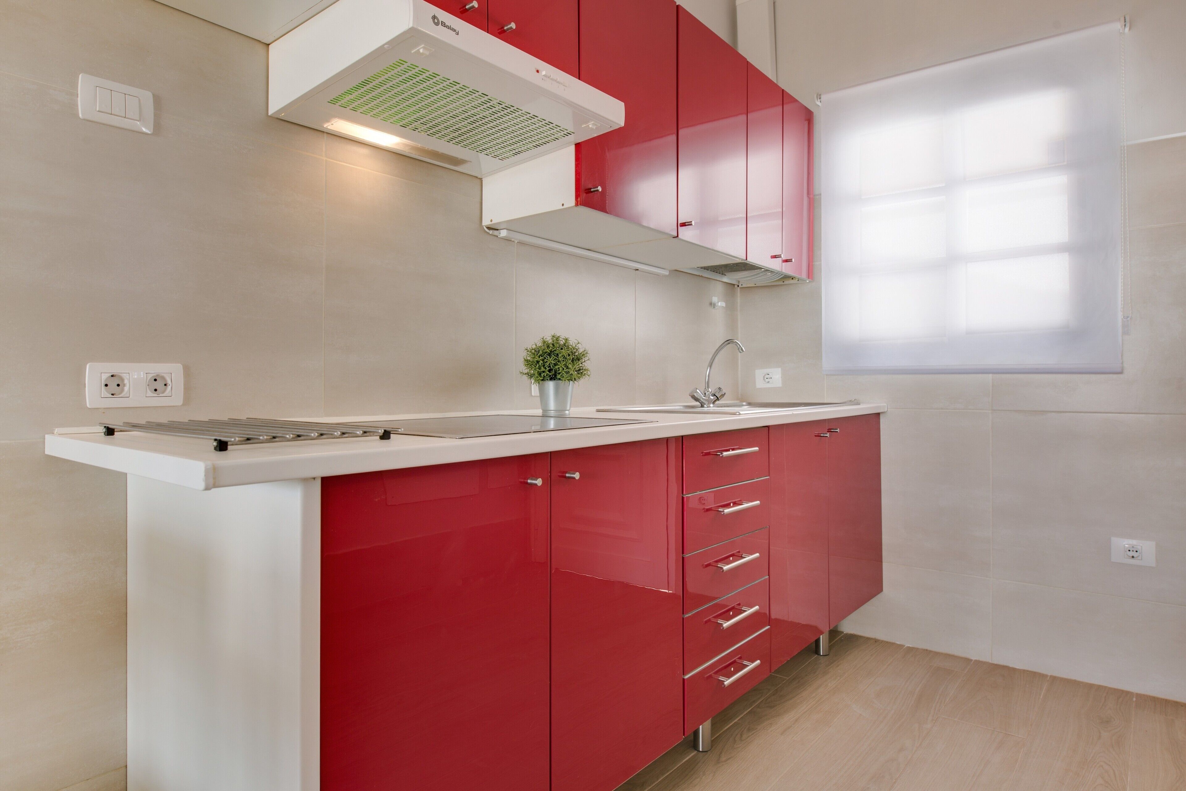 Une cuisine avec des meubles de couleur rouge, un plan de travail blanc, une petite plante et une fenêtre avec des stores.