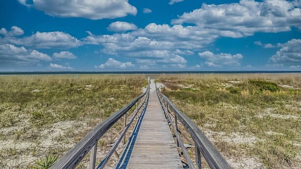 A wooden boardwalk leading through a grassy area with a clear blue sky and clouds above.