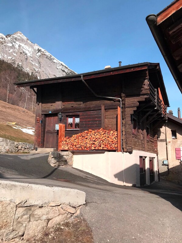 Un edificio rústico de madera con techo inclinado, una pila de leña al frente y un muro de piedra a la izquierda.
