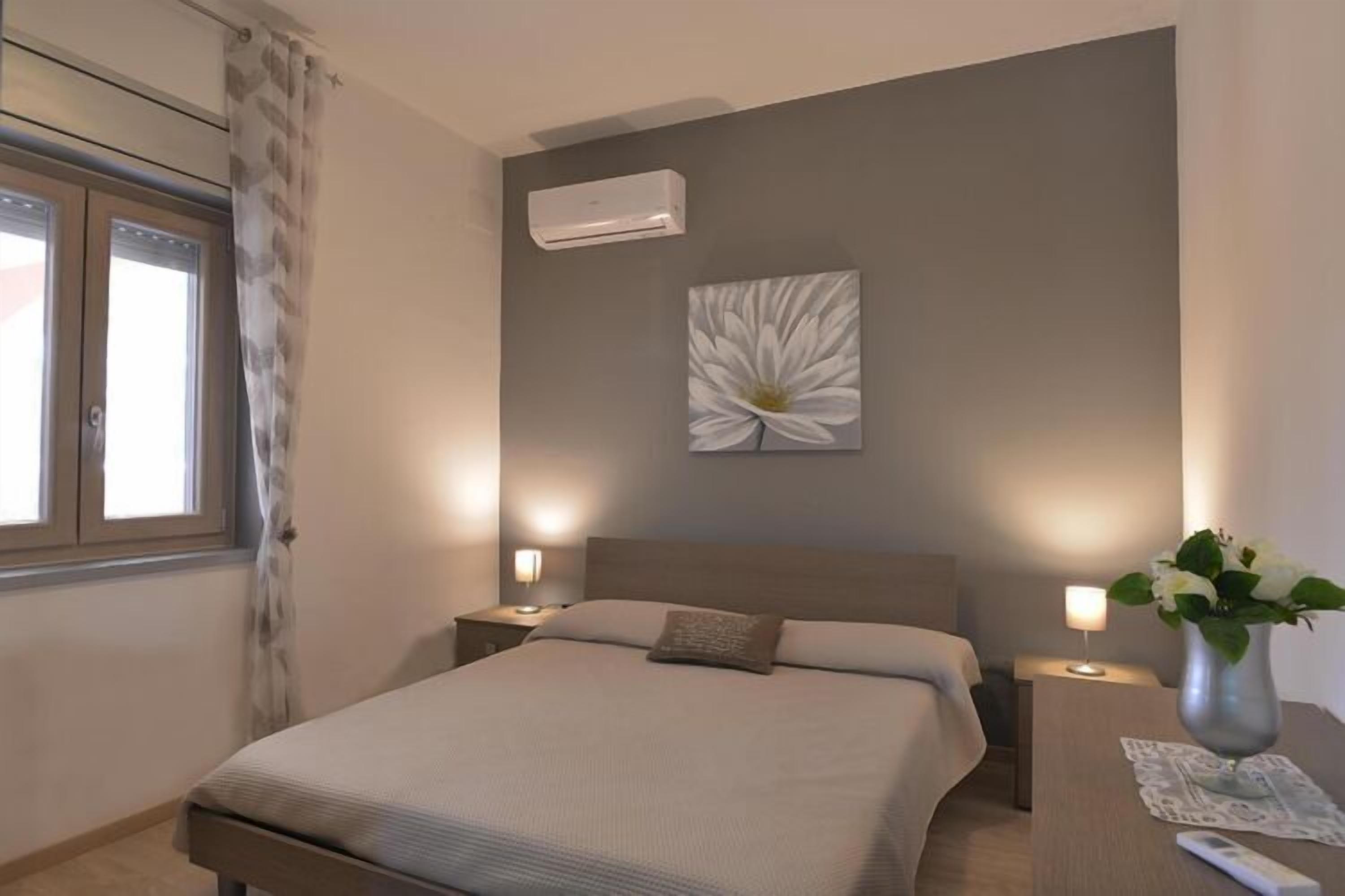 A bedroom with a bed, nightstands, an air conditioner, a wall-mounted artwork, and a vase with flowers.