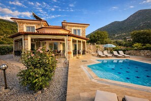 A two-story stone house with a swimming pool, outdoor seating, and a mountainous backdrop.