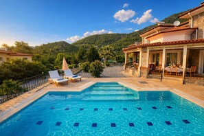 A swimming pool with lounge chairs, a covered patio with outdoor seating, and a mountainous backdrop.