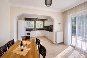 Un comedor con una mesa de madera, sillas y una botella de vino. Se ve una cocina al fondo con armarios blancos y una ventana.