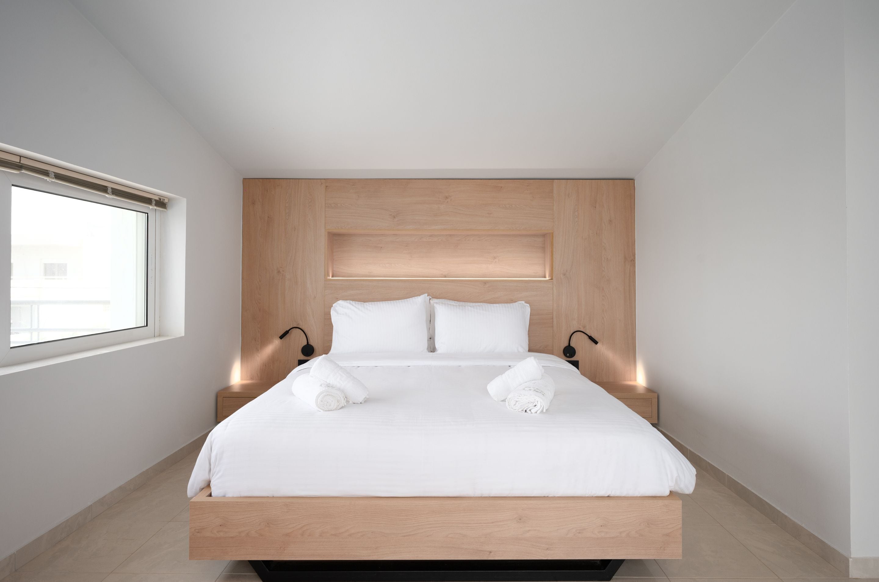 Una habitación de hotel moderna con cabecera de madera, una cama grande, dos mesitas de noche con lámparas y un ventanal que deja entrar luz natural.