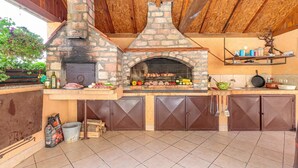 A stone-built outdoor kitchen with a brick oven, wooden cabinets, and a hanging shelf. - Pool & Sauna Countryside Retreat Klara (Velika Gorica)