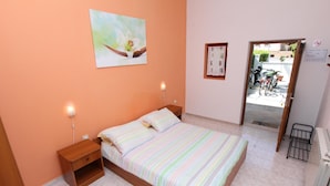 A bedroom with a bed, nightstands, a wall-mounted lamp, a radiator, and a door leading to an outdoor area with bicycles. - Apartments Denise (Rovinj)