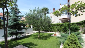 A well-maintained garden with various trees and shrubs, a clear pathway, and a building with a red-tiled roof in the background. - Apartments Denise (Rovinj)