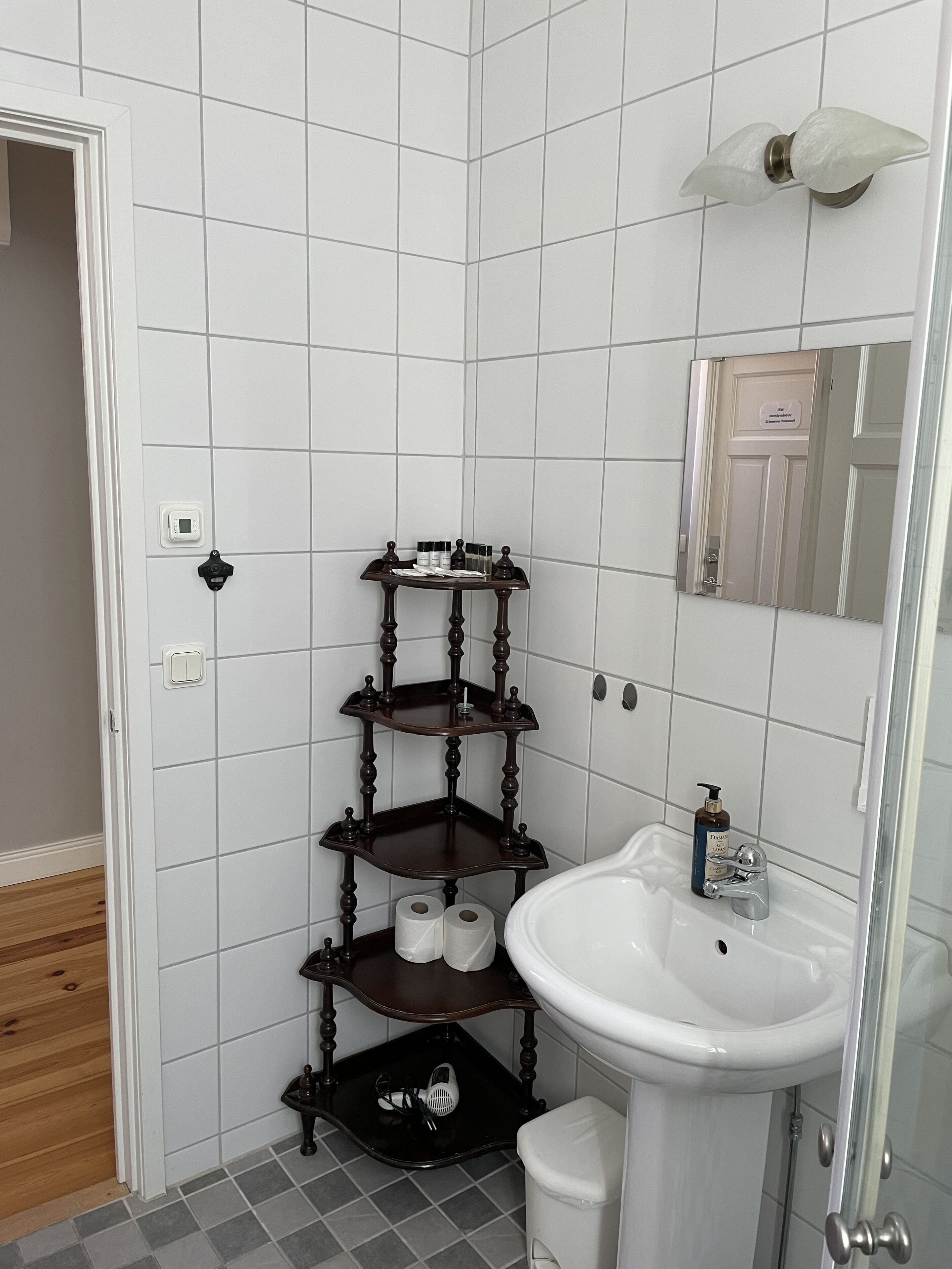 Une salle de bain avec un mur recouvert de carreaux blancs, un plancher en bois, un lavabo blanc, un miroir, un porte-serviettes et une étagère contenant des articles de toilette.