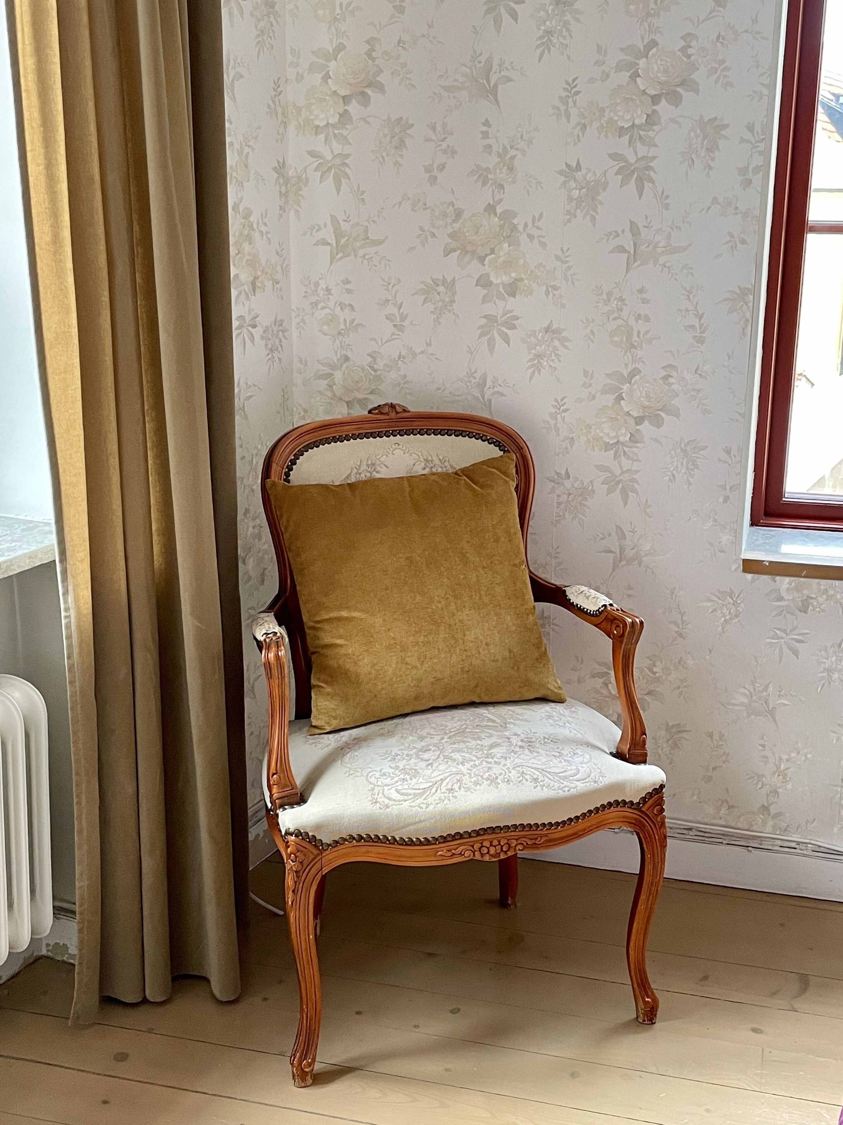 Ein Stuhl im Vintage-Stil mit Sitzkissen, platziert in einem Zimmer mit floralem Wandpapier und einem Fenster.