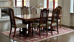 Une salle à manger avec une table en bois et six chaises, une cuisine à l’arrière-plan et une grande fenêtre donnant sur un immeuble.