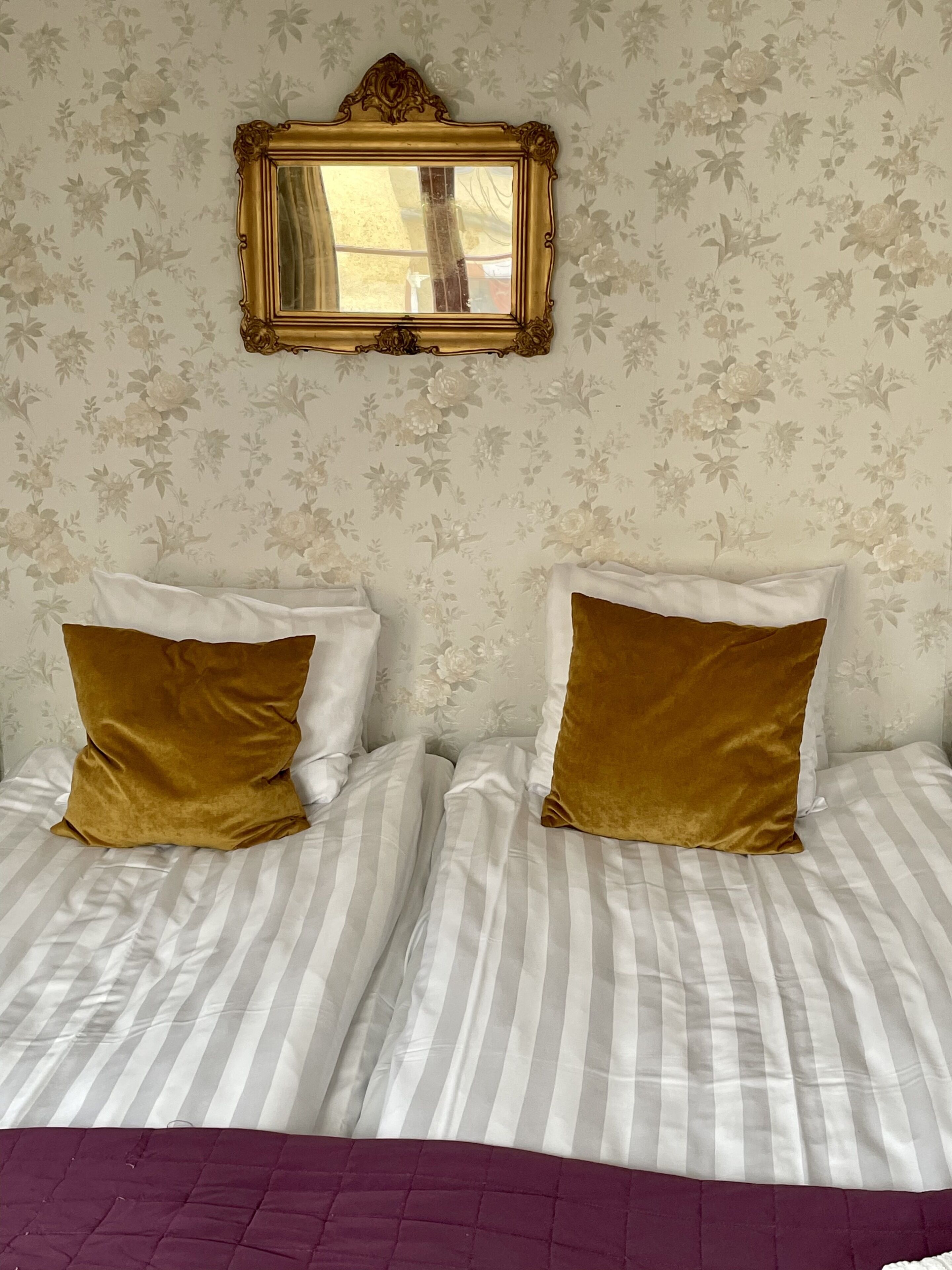 Una cama doble con ropa de cama a rayas y dos almohadas doradas, un espejo con marco dorado arriba y papel tapiz floral.