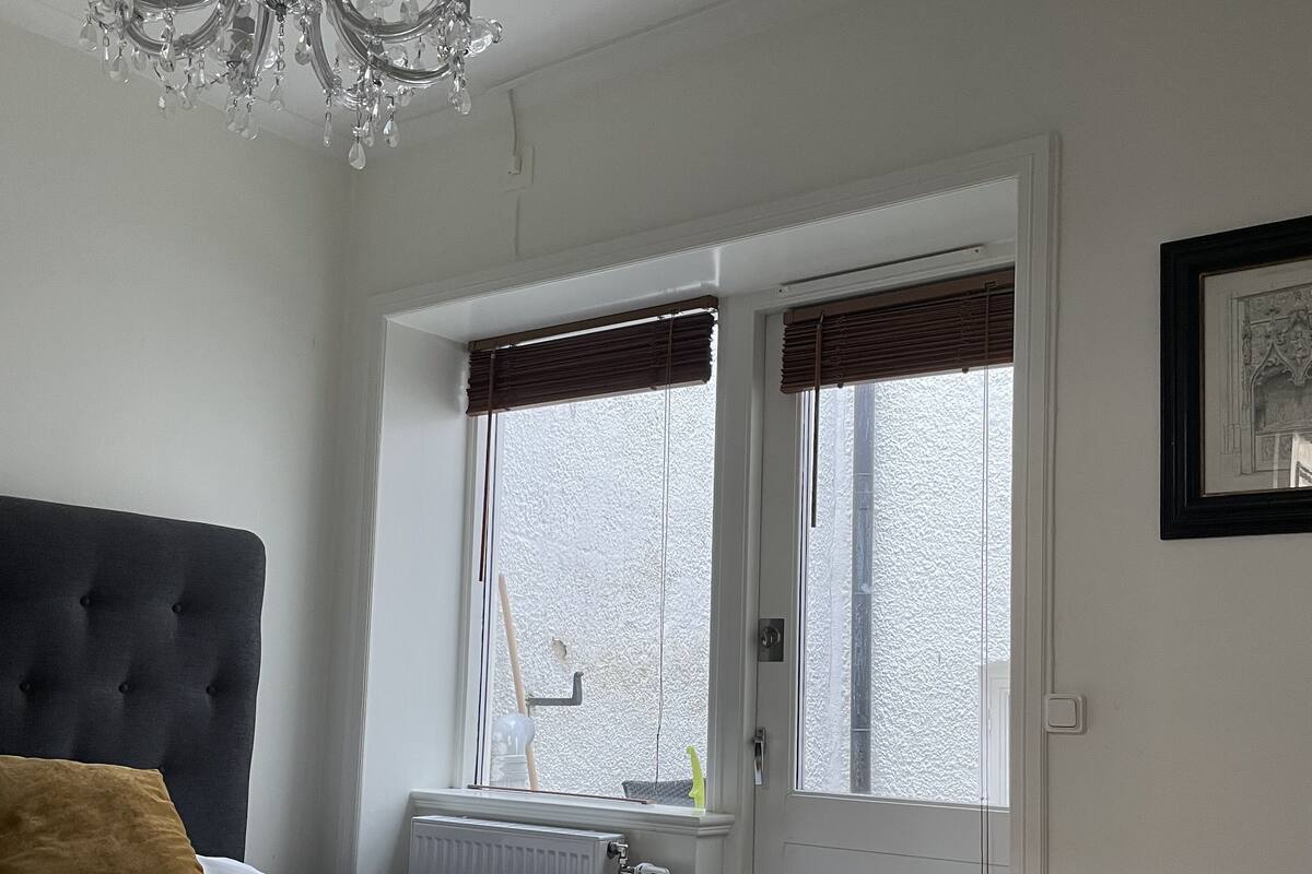 A bedroom with a bed, a chandelier, a radiator, and a window with blinds.