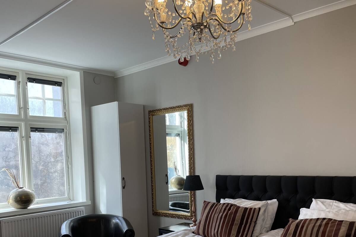 A hotel room with a large bed, a chandelier, a mirror, and a window with blinds.
