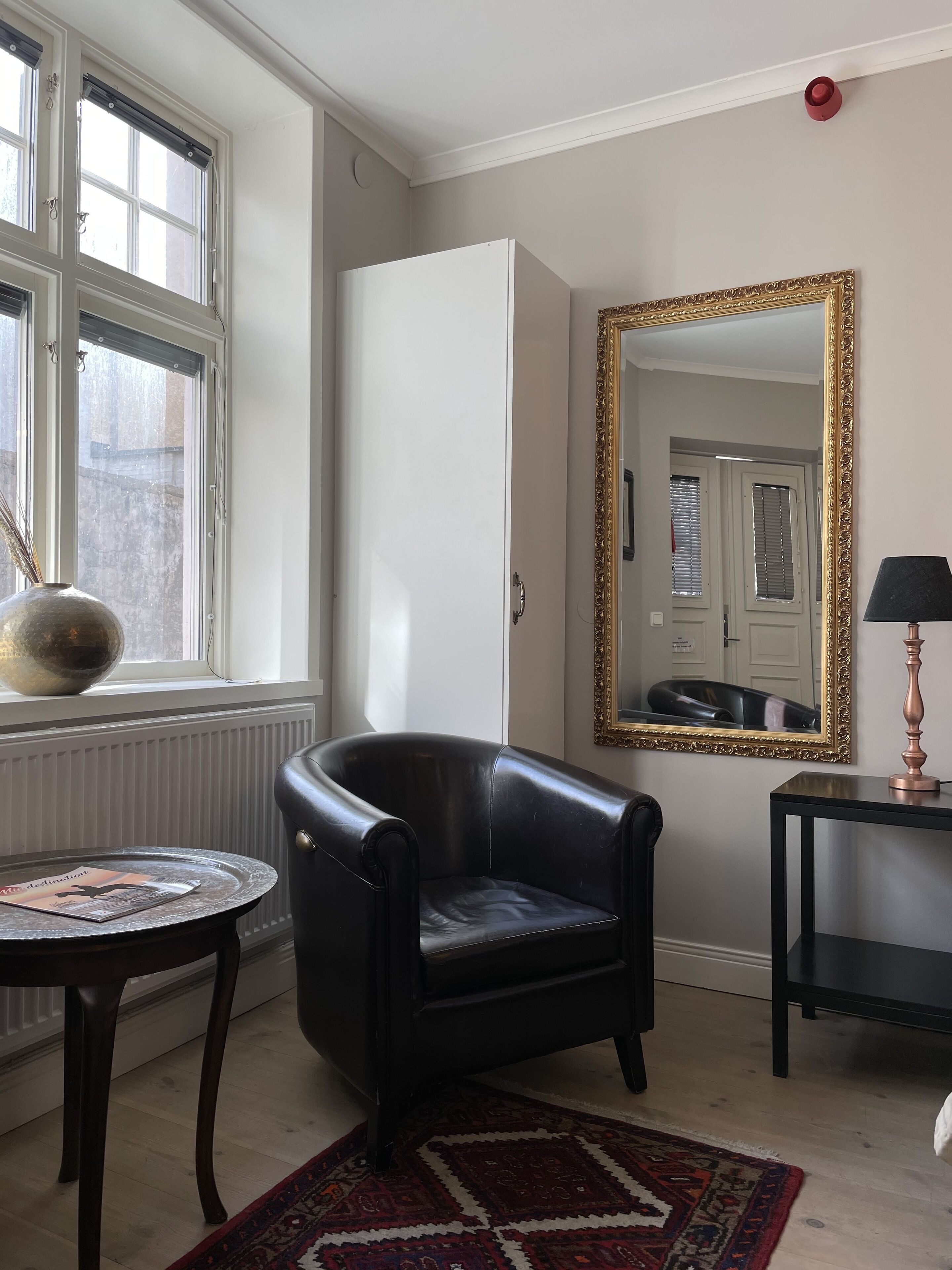 A room with a black leather chair, a round table with a magazine, a mirror with a gold frame, and a radiator.