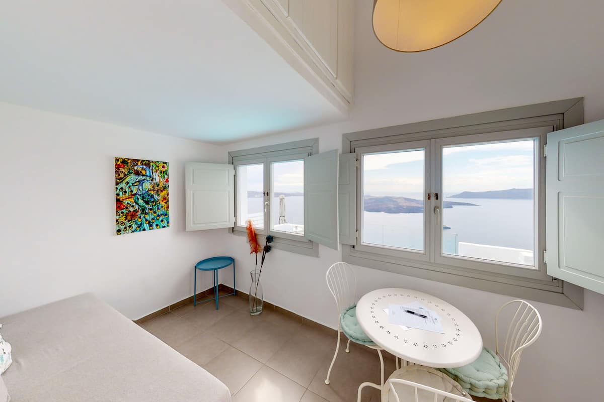 A room with a bed, a dining table with chairs, a vase with flowers, a painting, and a window with a view of the sea.