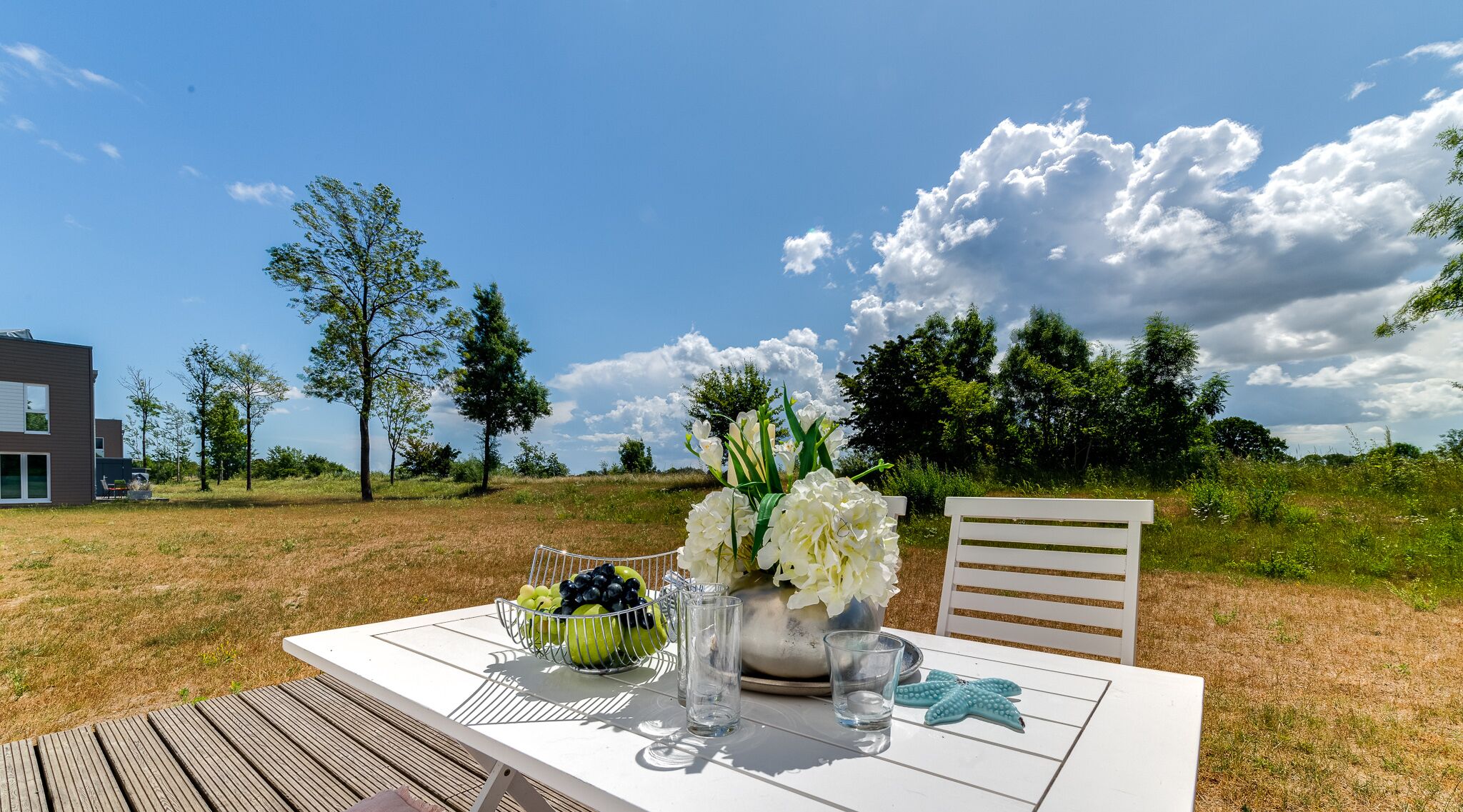 An outdoor dining table with a white tablecloth, a bowl of fruit, a vase of flowers, and a glass of water.