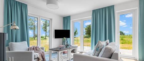 A modern living room with a flat-screen TV, two sofas, a coffee table, and large windows with teal curtains.
