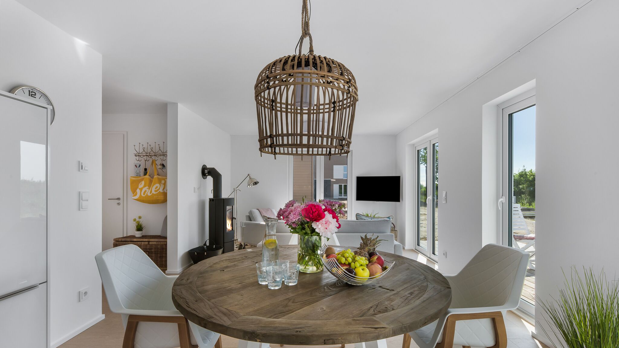 A modern dining area with a round wooden table, white chairs, a wicker pendant light, and a vase with flowers.