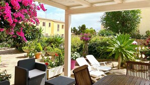 Un espace extérieur aménagé, abrité et doté de chaises en osier et d’une table, entouré de plantes en pot et de fleurs aux couleurs vives.