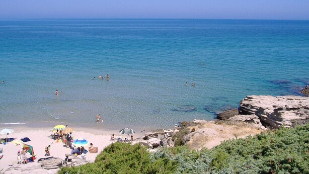 Une plage avec une eau d’un bleu limpide, des affleurements rocheux, et des gens qui se baignent et prennent des bains de soleil.