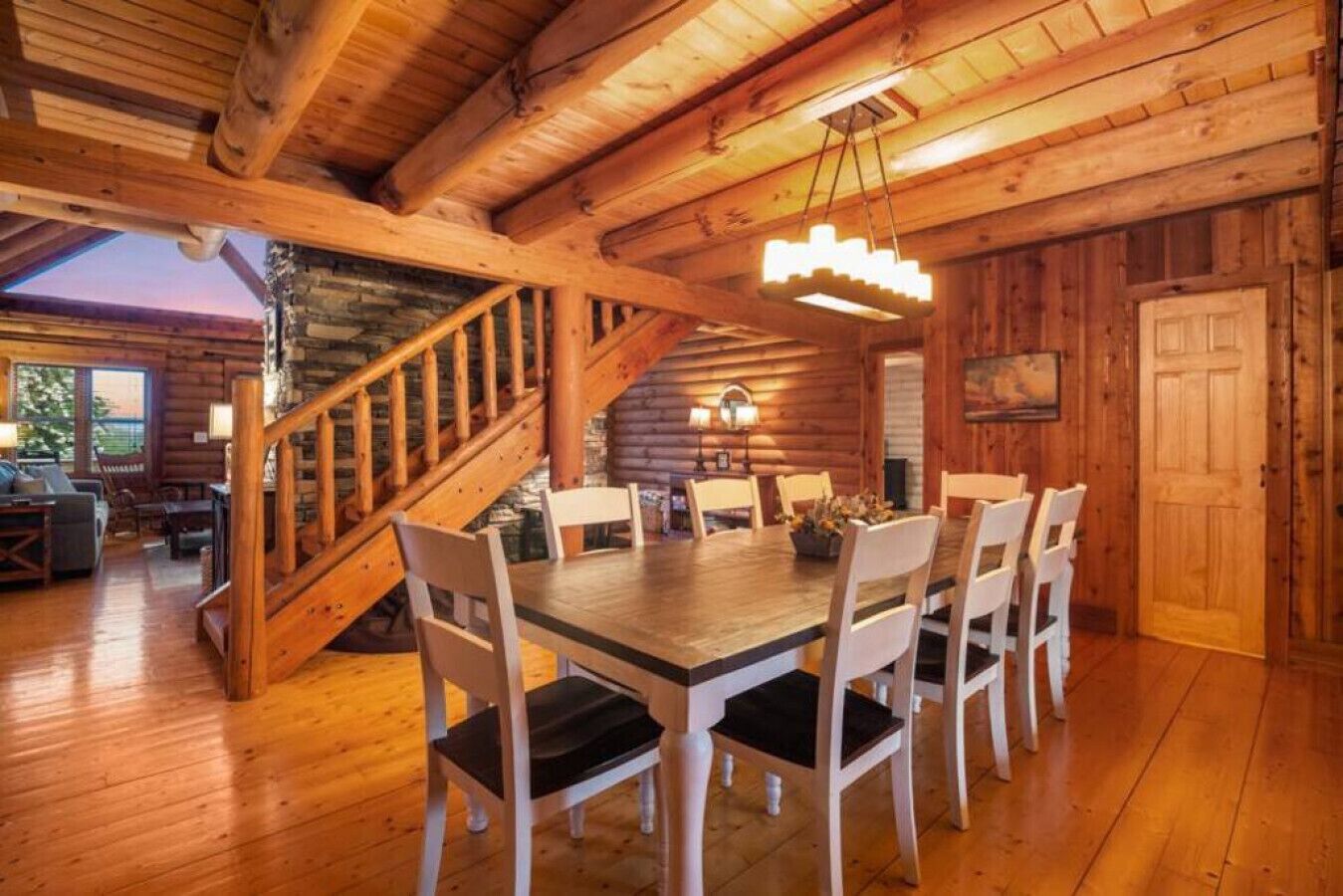 A wooden cabin interior with a dining table, chairs, a staircase, and a chandelier.