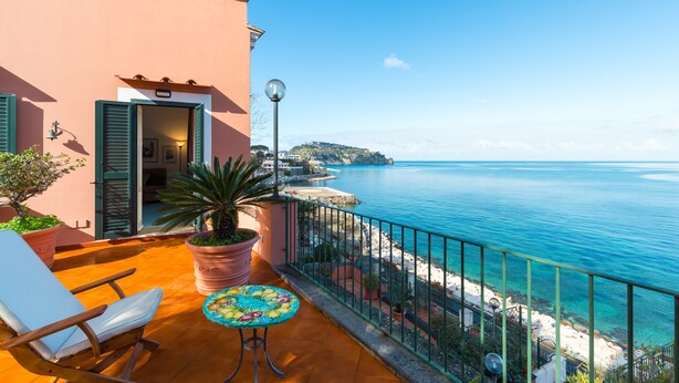 A balcony with a view of the sea, a chair, a table with a plant, and a building with a door and shutters. - Terrazza Morgera by Napoliapartments (Casamicciola Terme)