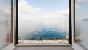 A view through a window of a coastal scene with clear water, a rocky shoreline, and a distant landmass. - Terrazza Morgera by Napoliapartments (Casamicciola Terme)