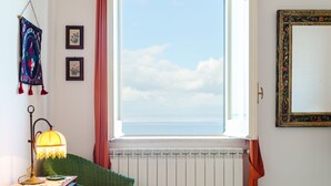 A bedroom with a large window, a bed with a pink and white striped bedspread, a green chair, a lamp, and a framed mirror on the wall. - Terrazza Morgera by Napoliapartments (Casamicciola Terme)