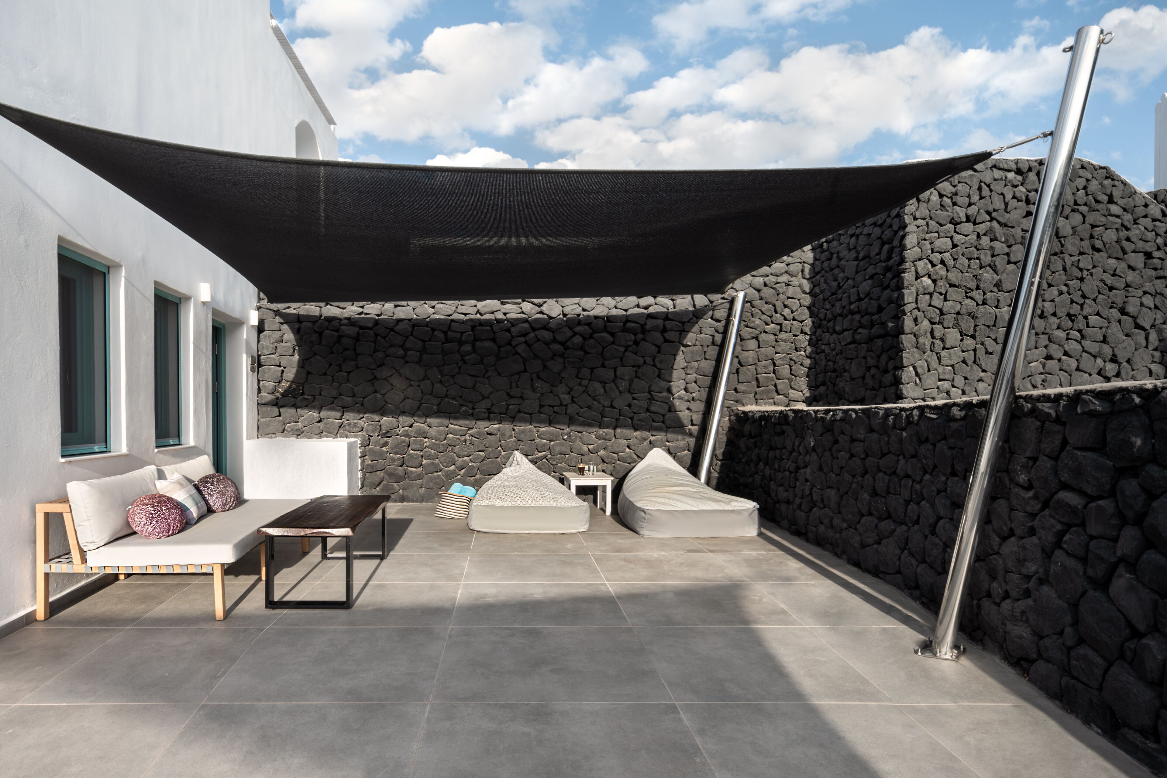 A modern outdoor patio with a stone wall, a white sofa, a wooden table, and a large shade sail.