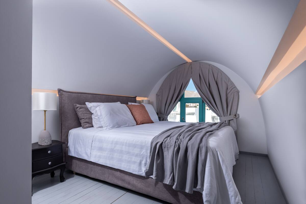 Une chambre avec un plafond en pente, un lit avec une literie blanche, une table de chevet avec une lampe et une fenêtre avec des rideaux.