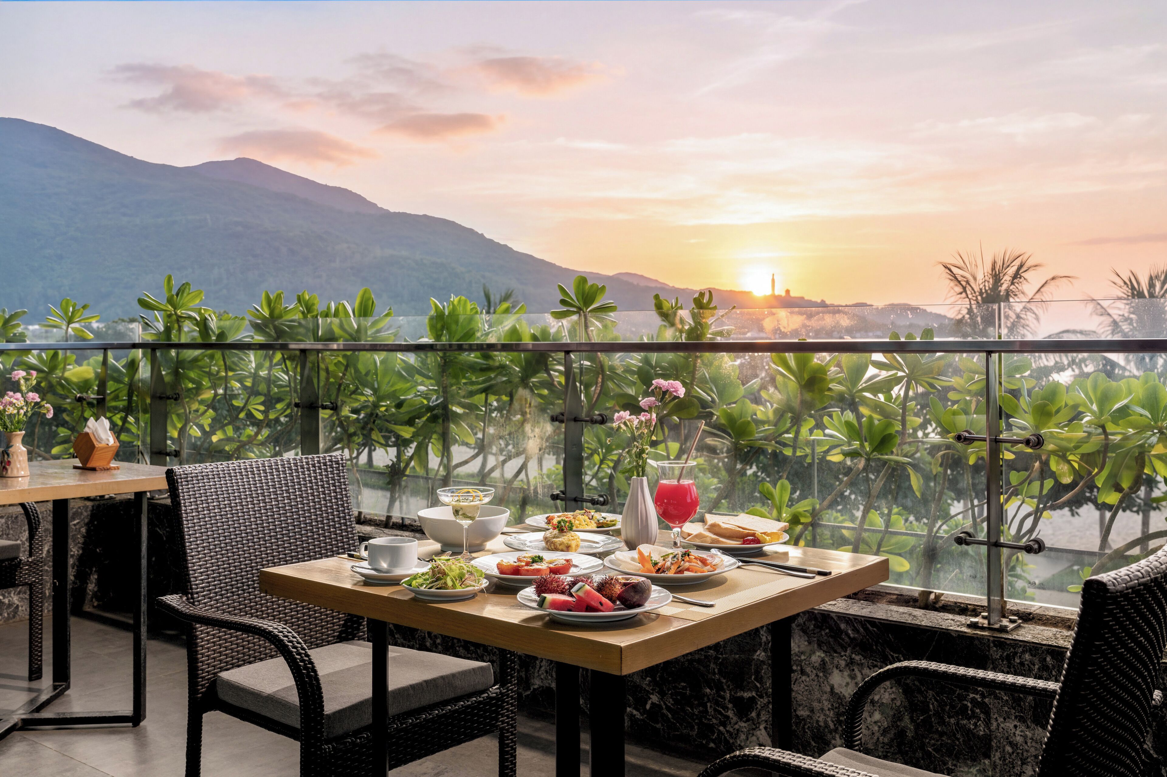 Une table à manger avec des assiettes remplies de nourriture, un verre d’une boisson rouge et un bol blanc, le tout sur fond de coucher de soleil et de montagnes.