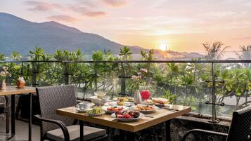 Une table à manger avec des assiettes remplies de nourriture, un verre d’une boisson rouge et un bol blanc, le tout sur fond de coucher de soleil et de montagnes.