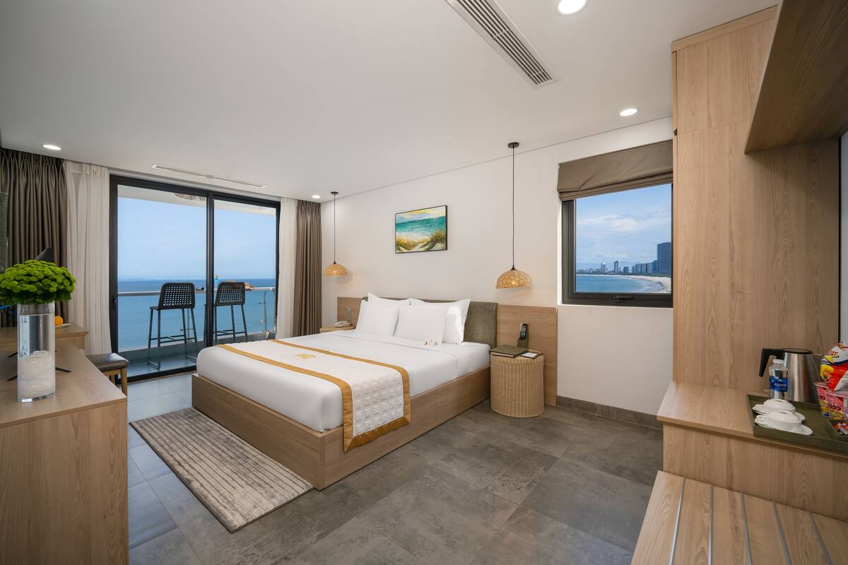 A modern hotel room with a large bed, a bedside table, a small table with a vase, a chair, and a view of the sea.