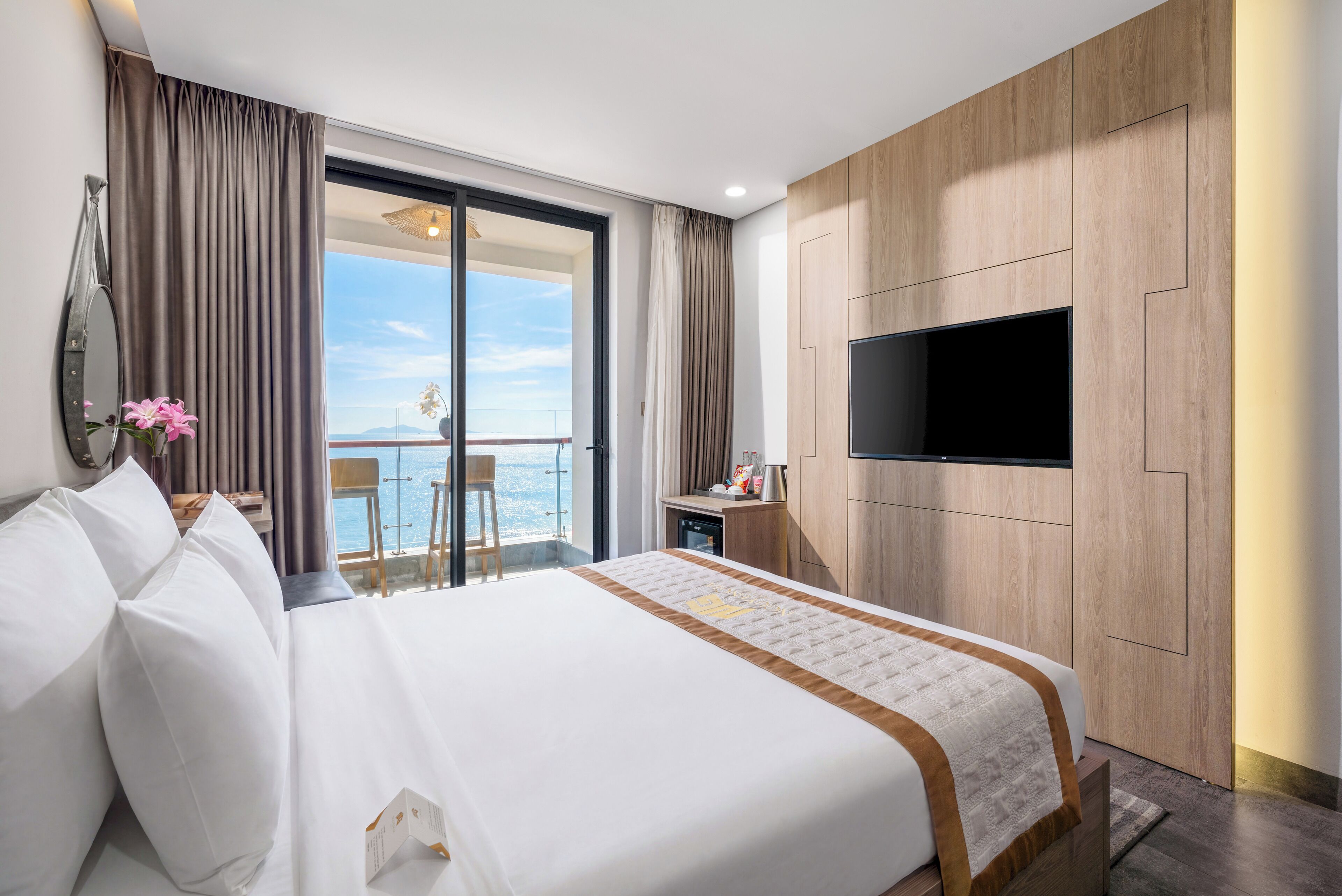 Habitación de hotel con una cama grande, un televisor de pantalla plana montado en la pared, un balcón con vistas y una pequeña mesa con una silla.