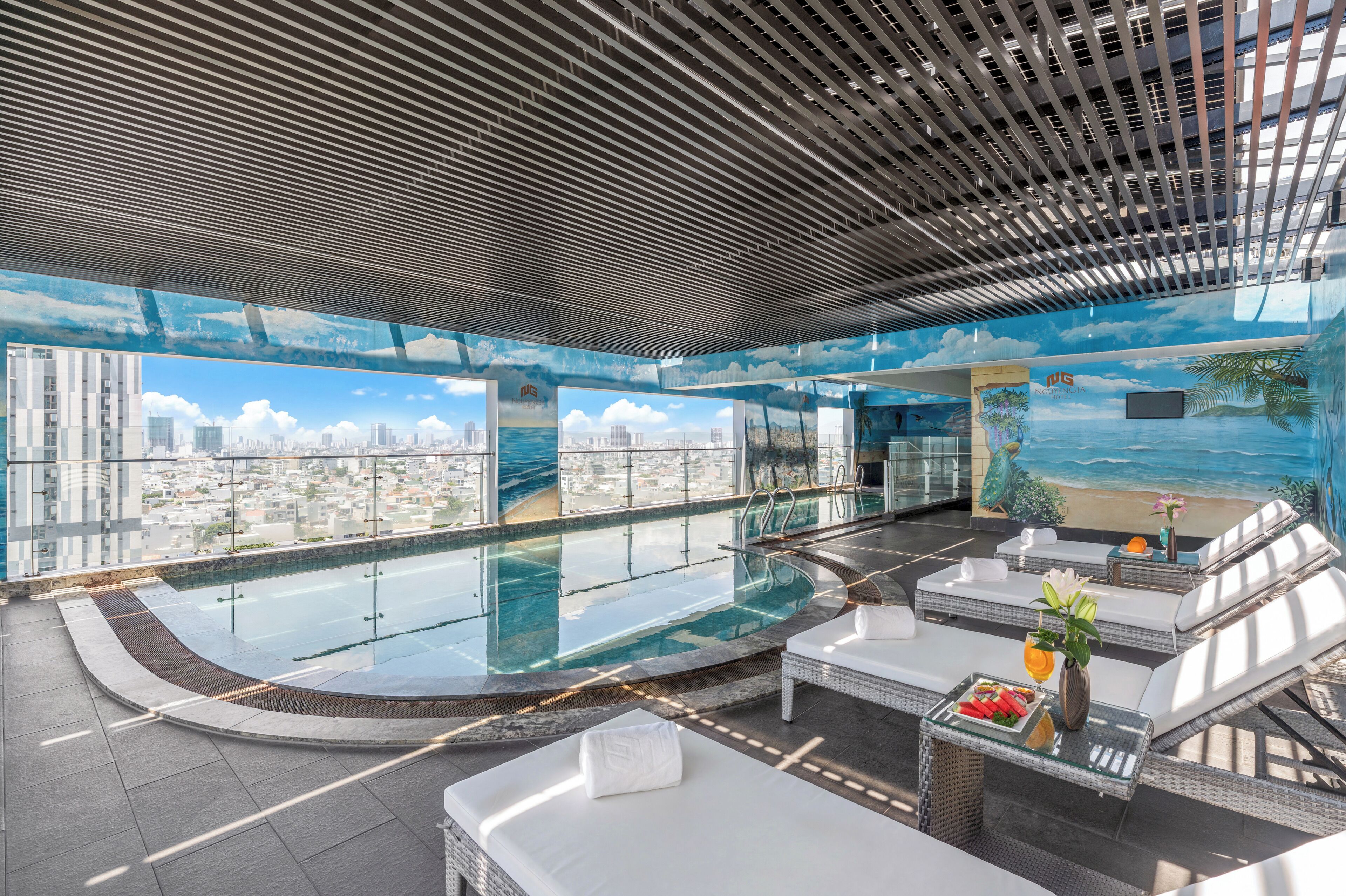 Una piscina infinita en la azotea con vista a la ciudad, zona de descanso con asientos y un mural de una escena de playa.