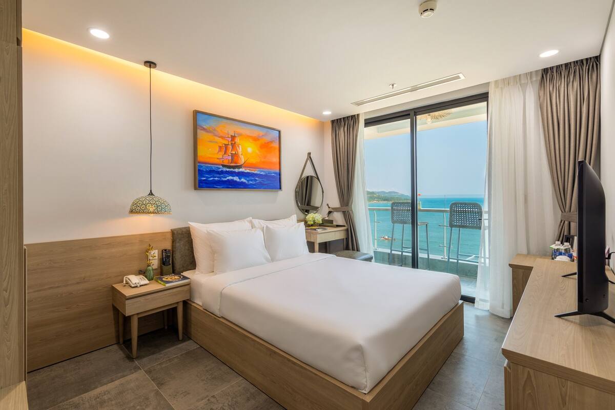 A hotel room with a large bed, a desk with a computer, a phone, and a painting of a ship on the wall.
