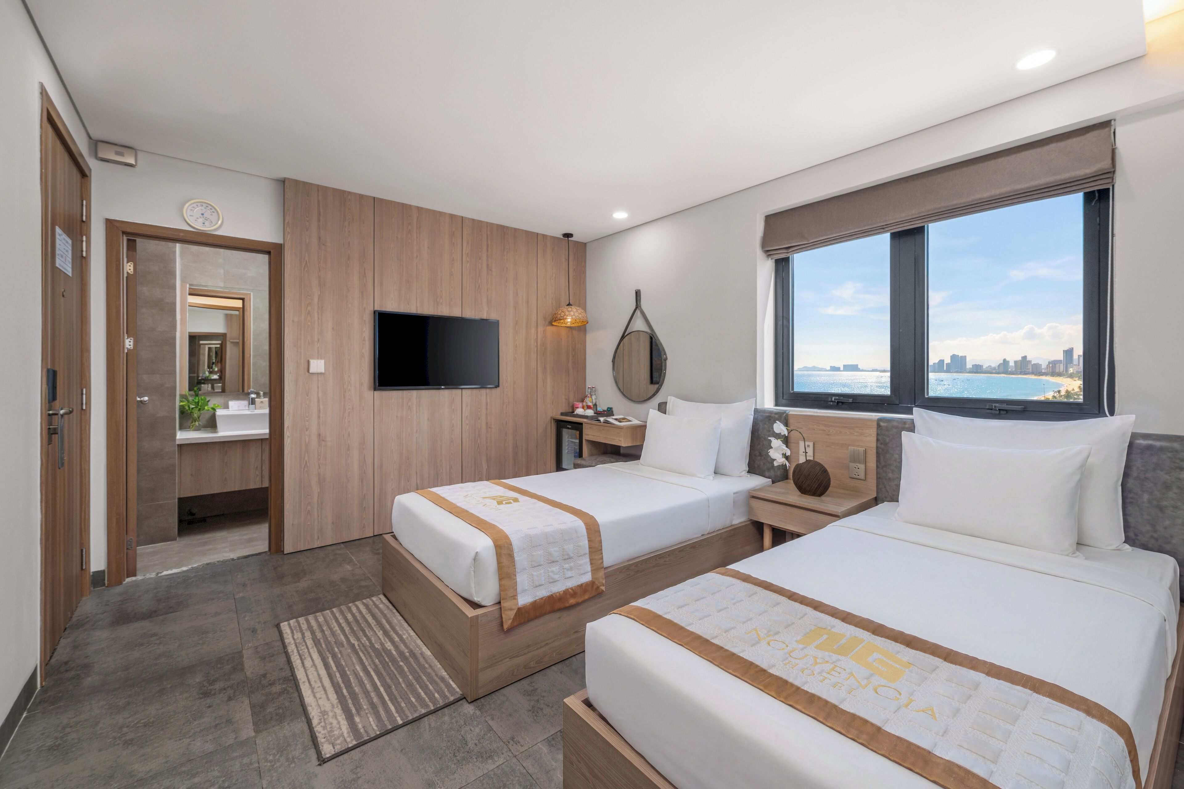 A hotel room with two beds, a TV, a window with a city view, and a bathroom with a mirror and sink.