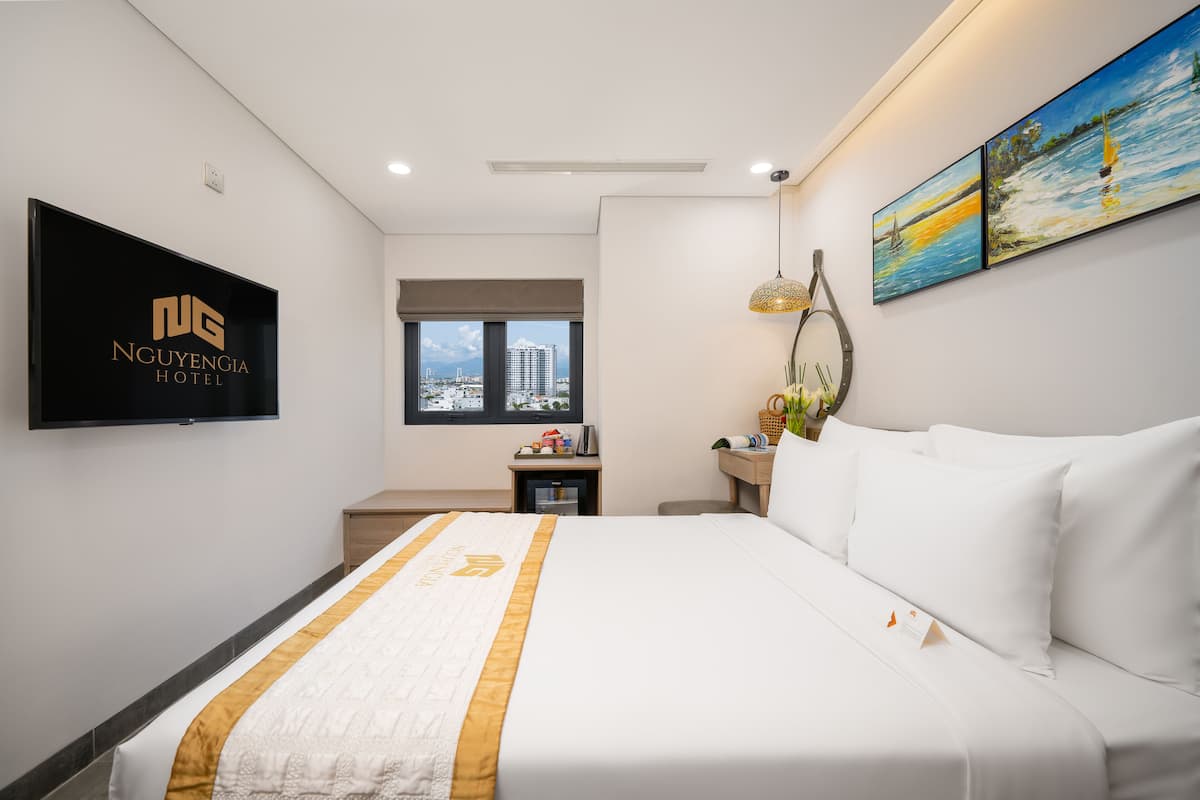 A hotel room with a large bed, a flat-screen TV mounted on the wall, a window with a city view, and artwork on the wall.