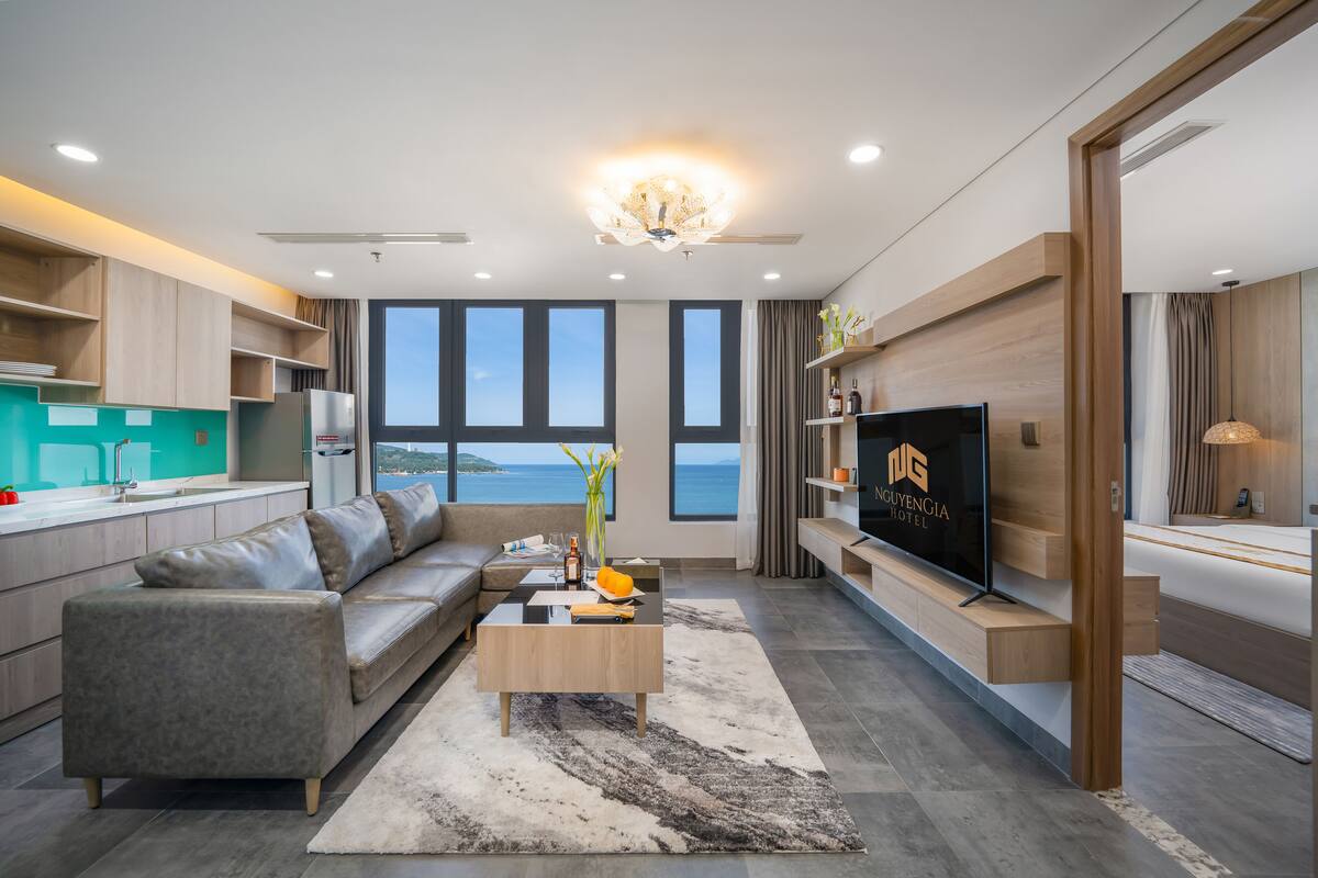 A modern living room with a sofa, coffee table, and TV. It features large windows with a view of the sea.